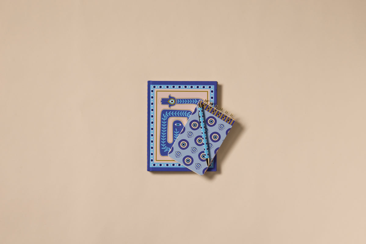 A Dopo Eye Journals in blue and white, with geometric and abstract designs, rest on a beige surface; the lay-flat notebooks make a thoughtful gift for thinkers.