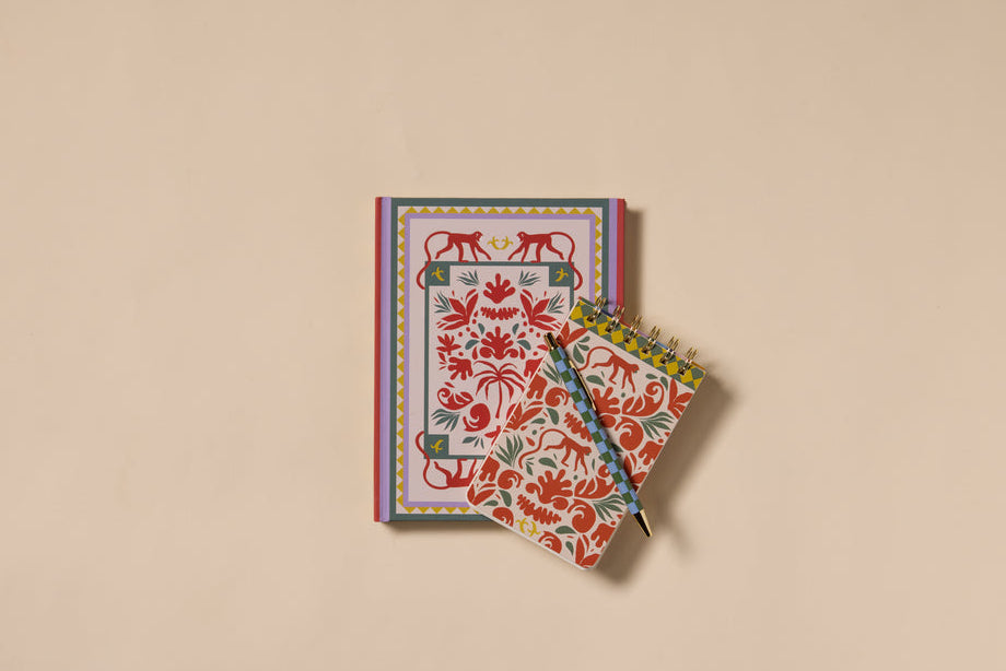 The A Dopo Monkey Twin Wire Notepad, featuring playful hand-drawn animal and floral designs, sits on a beige surface beside a larger closed notebook, with a pen resting on the spiral notepad.