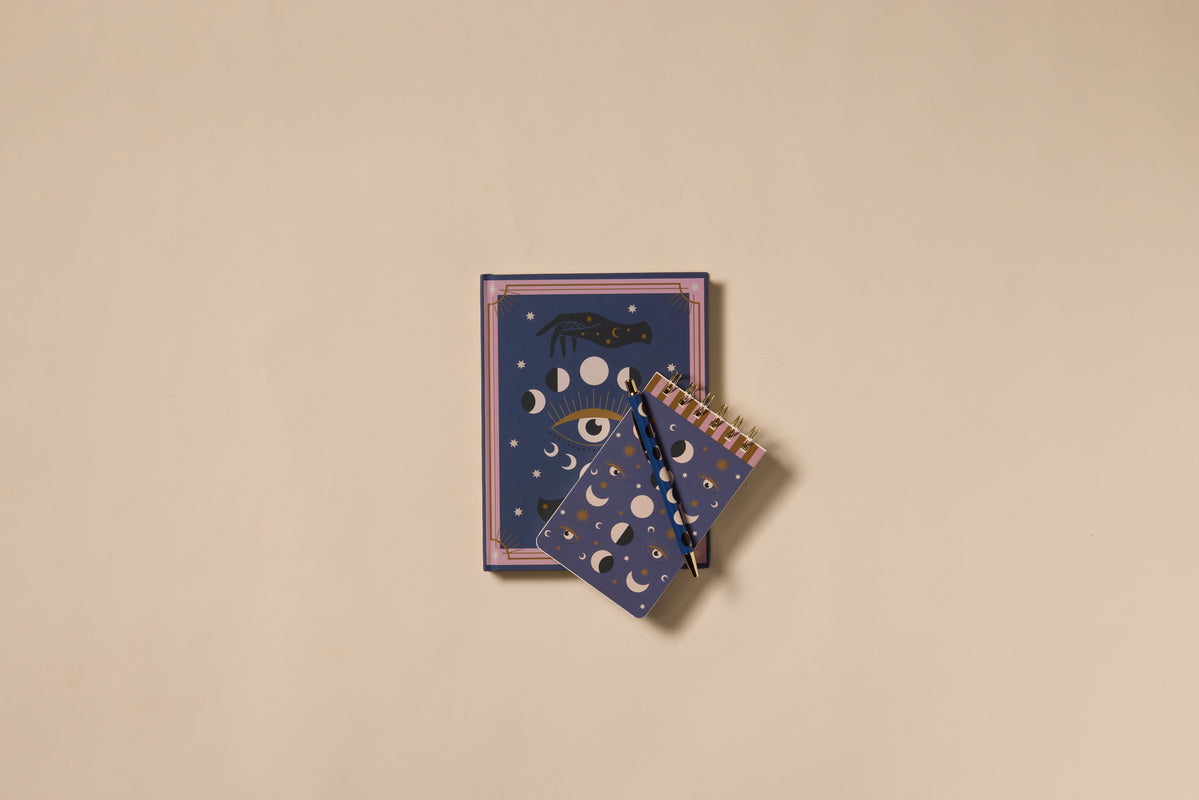 Two blue A Dopo Journals - Moon Phases, featuring gold moon phases, an eye, and stars on the covers, are placed on a beige background. One journal is larger, while the other is a spiral-bound version with two pencils resting on it.