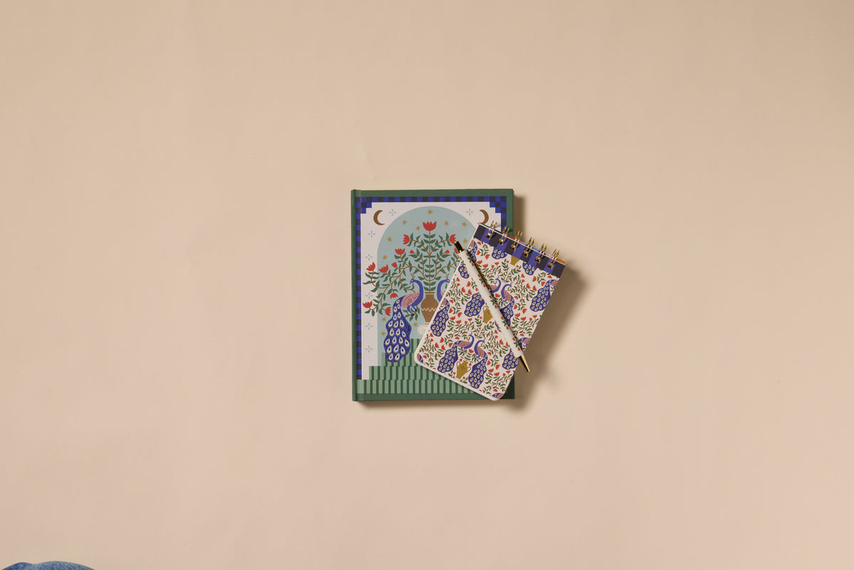 Two A Dopo Peacock Journals with vibrant floral and tree designs, featuring soft-touch covers—one spiral-bound—are stacked on a plain beige surface, with the spiral-bound notebook positioned diagonally on top.