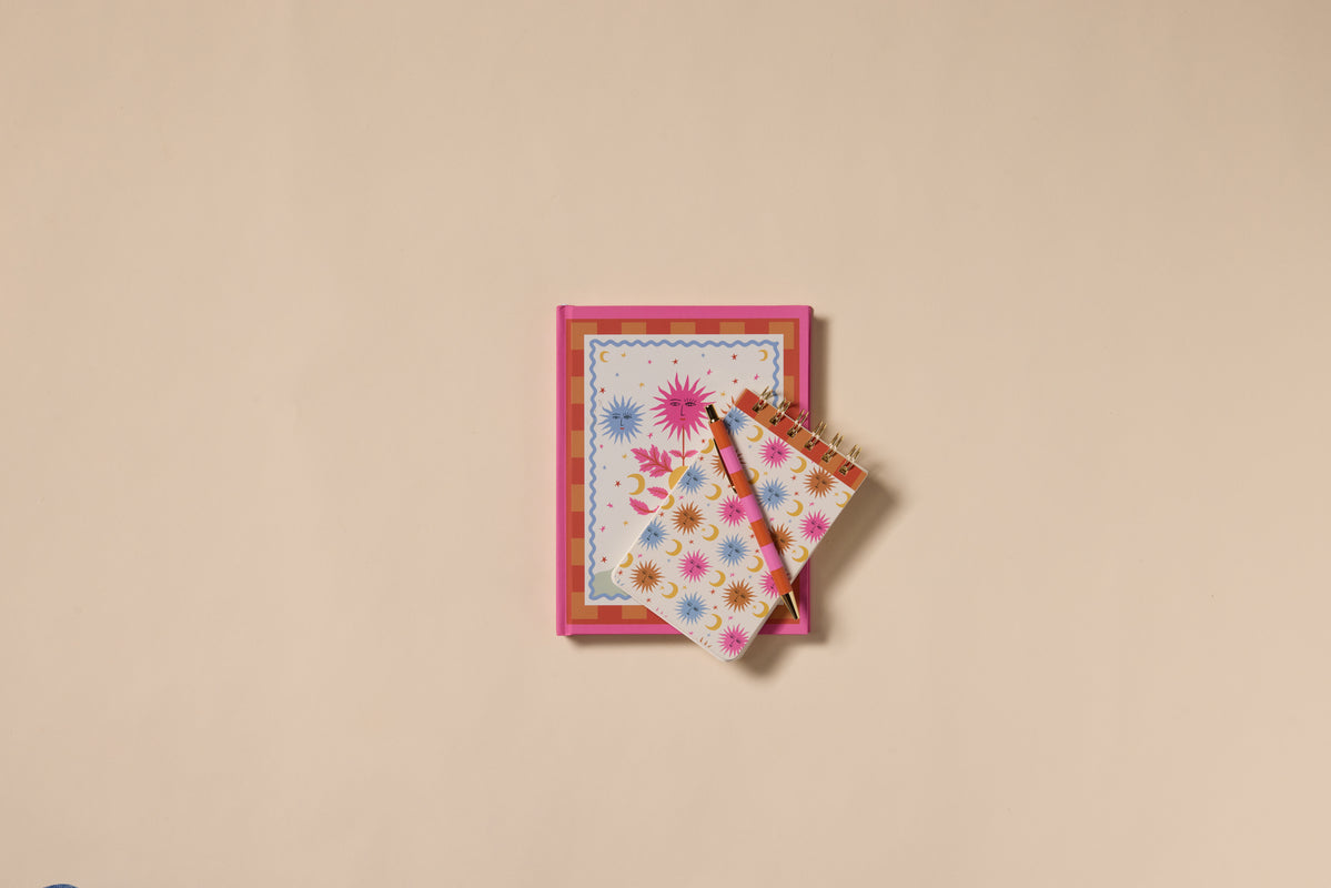 A Dopo Sun Twin Wire Notepad with a colorful, starburst cover rests on a beige surface. On top is a matching notepad and pen featuring vibrant hand-drawn Mediterranean designs, creating a coordinated stationery set.