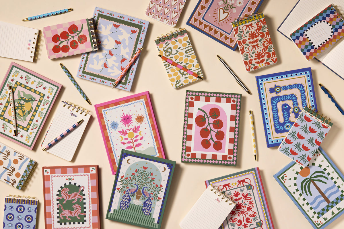 Colorful notebooks and notepads with various decorative covers are arranged on a beige surface, accompanied by pens in matching patterns.