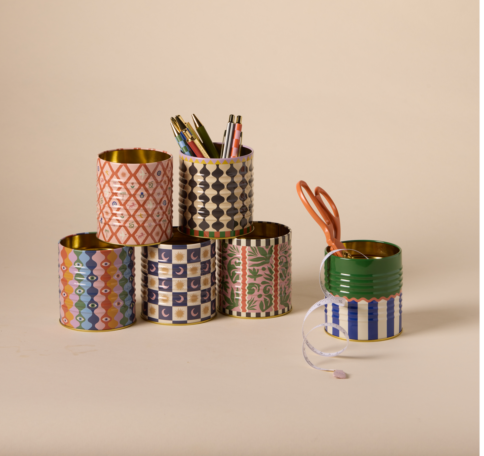 Six patterned tin cans arranged in a pyramid, some holding pens, pencils, and scissors, against a plain beige background.