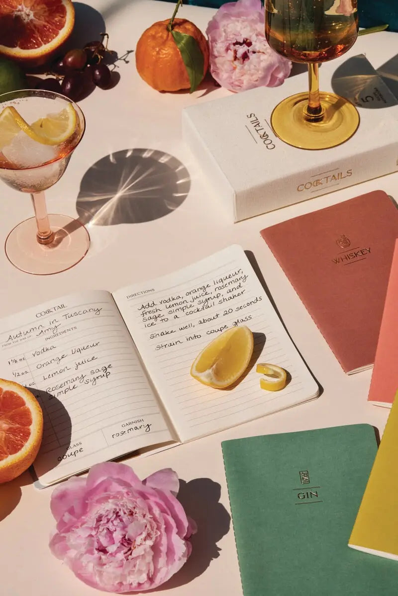 The Cocktail Recipe Box Set, open cloth bound notebooks with cocktail recipes, assorted fruits, a wine glass, and pink peonies are elegantly arranged on a beige surface in natural light. A handy conversion table is also visible.