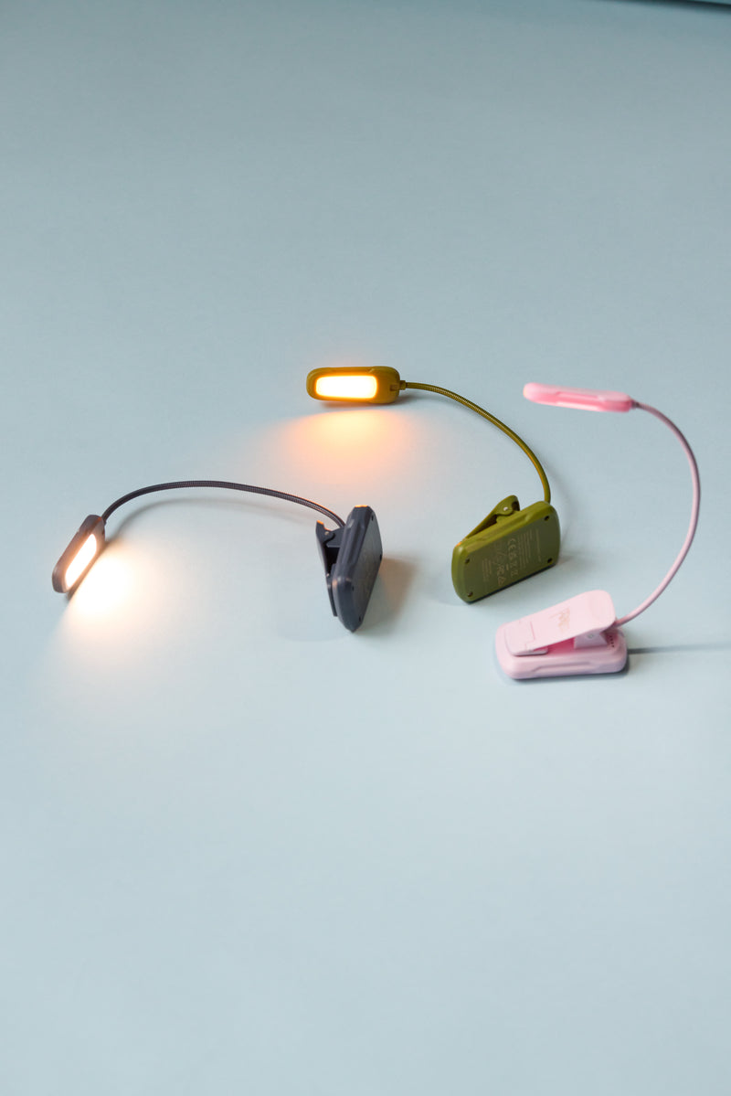 A Book Light - Navy with a flexible neck is shown on a light blue surface, its LED lit and casting a warm glow—perfect as a portable reading light or travel booklight.