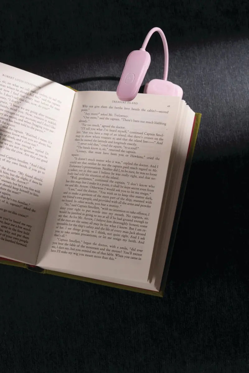 An open book rests on a dark surface, illuminated by the compact and lightweight Book Light - Lilac with 9 LED settings attached to the top.