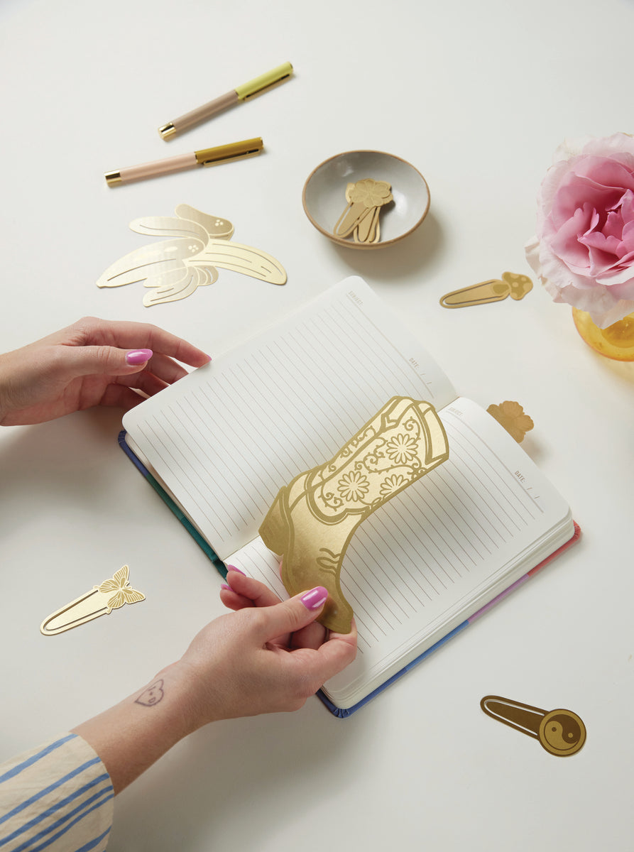 A person places a large gold cowboy boot bookmark on an open lined notebook. Brass Page Tabs - Mushrooms + Butterfly, pens, and a pink flower in a vase are also arranged on the white table.
