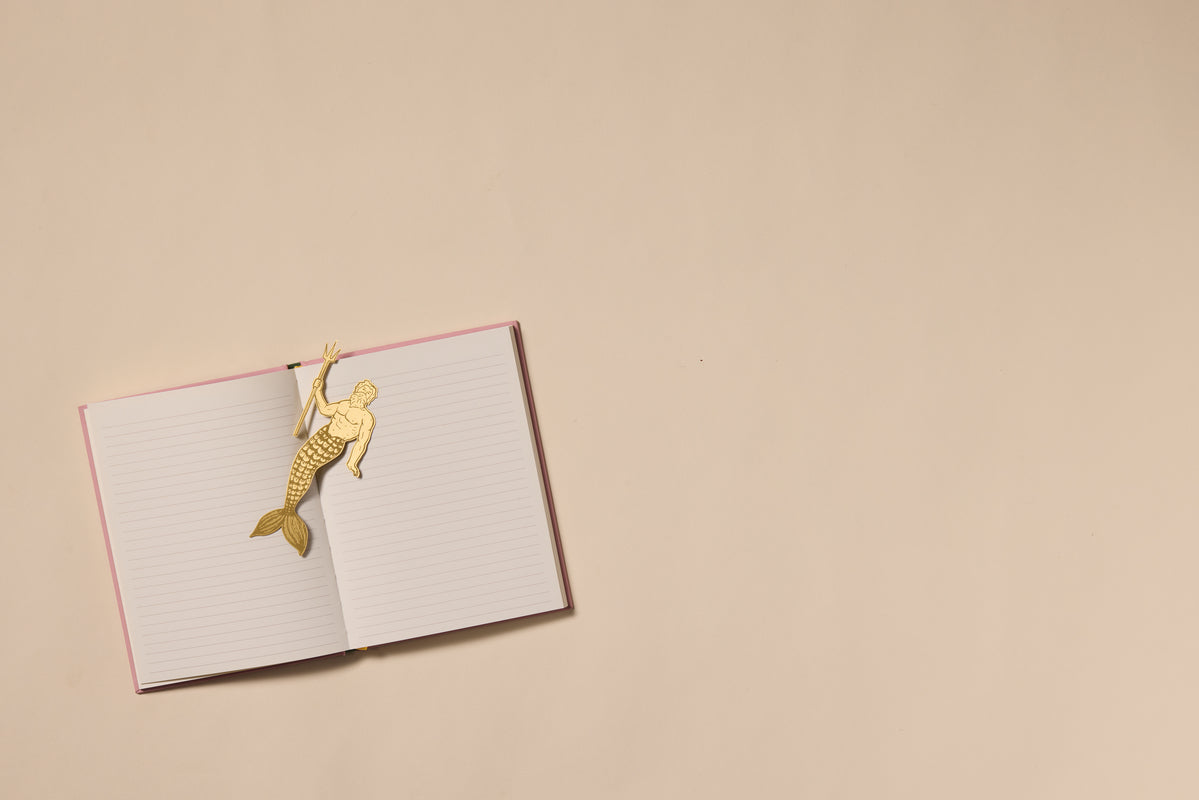An open pink-covered lined notebook rests on a beige surface with the Brass Bookmark - Poseidon, shaped like a mermaid, clipped to the top left page. The notebook is positioned to the left, leaving empty space on the right.