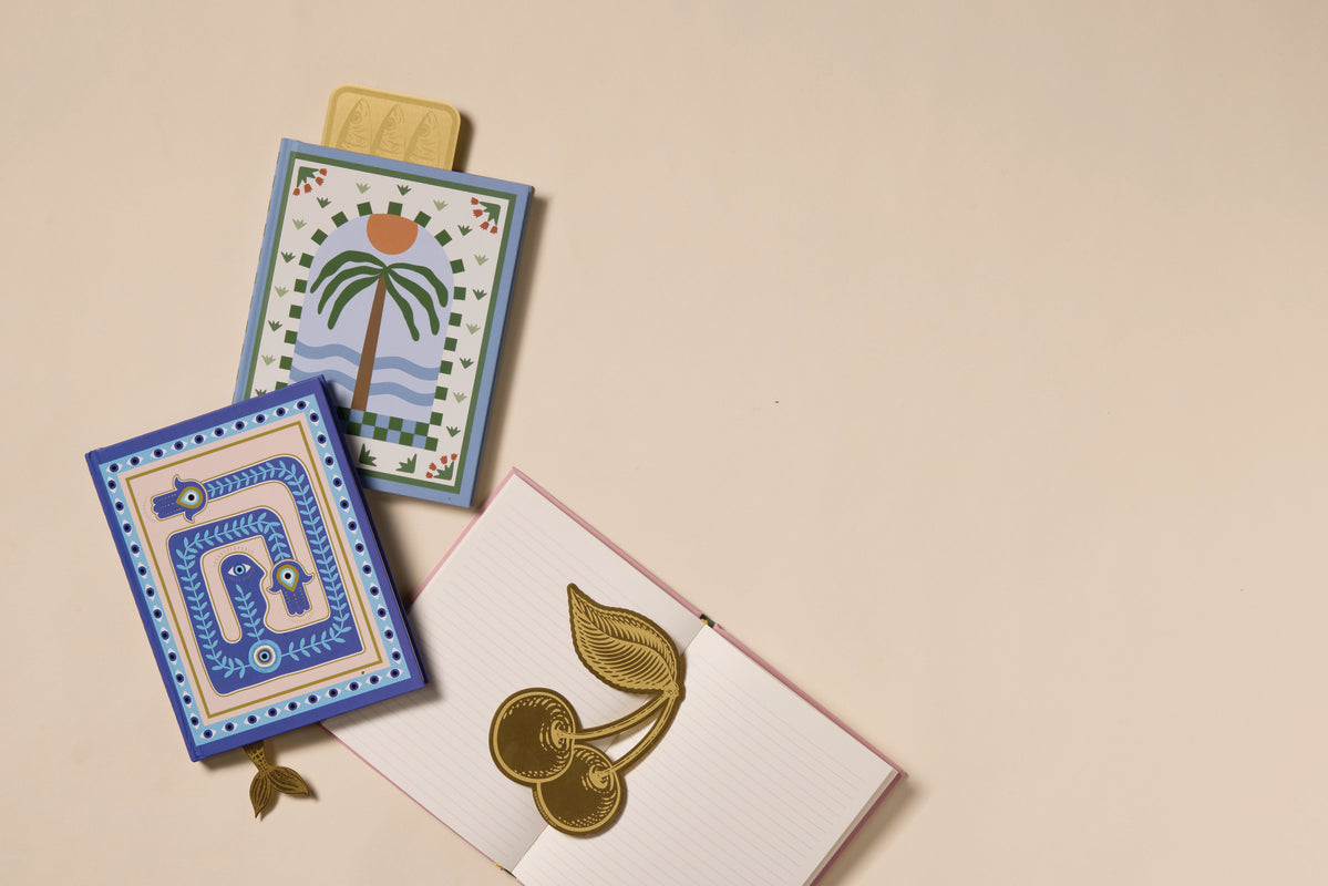 Two decorative notepads—one with a palm tree and sun, the other with blue patterns—are placed beside an open lined notebook adorned with the Brass Bookmark - Cherries, all set on a light beige surface.
