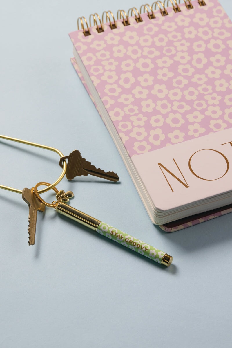 A Pen Keychain - Stay Groovy and a pair of gold keys rest beside a pink floral NOTES notebook, all arranged on a light blue surface—an ideal combo for any on-the-go scribe.
