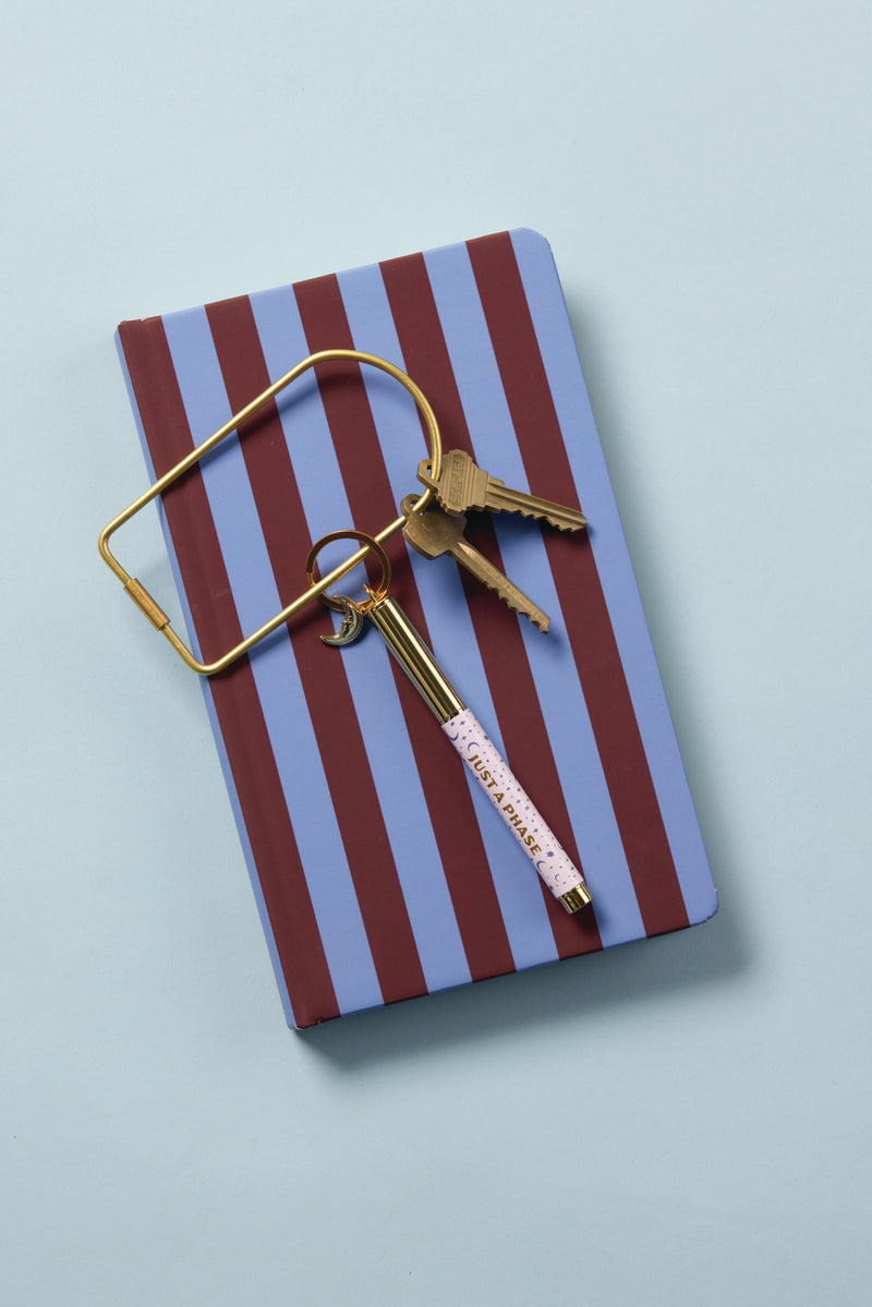 A Soft Touch Journal in Purple + Nutmeg Stripe rests on a light blue surface, topped with a gold key ring holding two keys and a gold IMPORTANT-labeled pen.