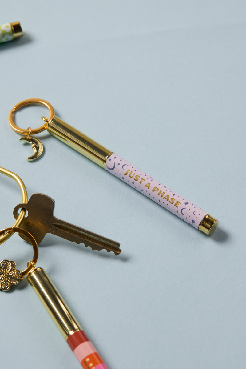 The Pen Keychain - Just a phase features a gold keychain with a key and a pastel mini ballpoint pen inscribed with "JUST A PHASE," displayed on a light blue surface.