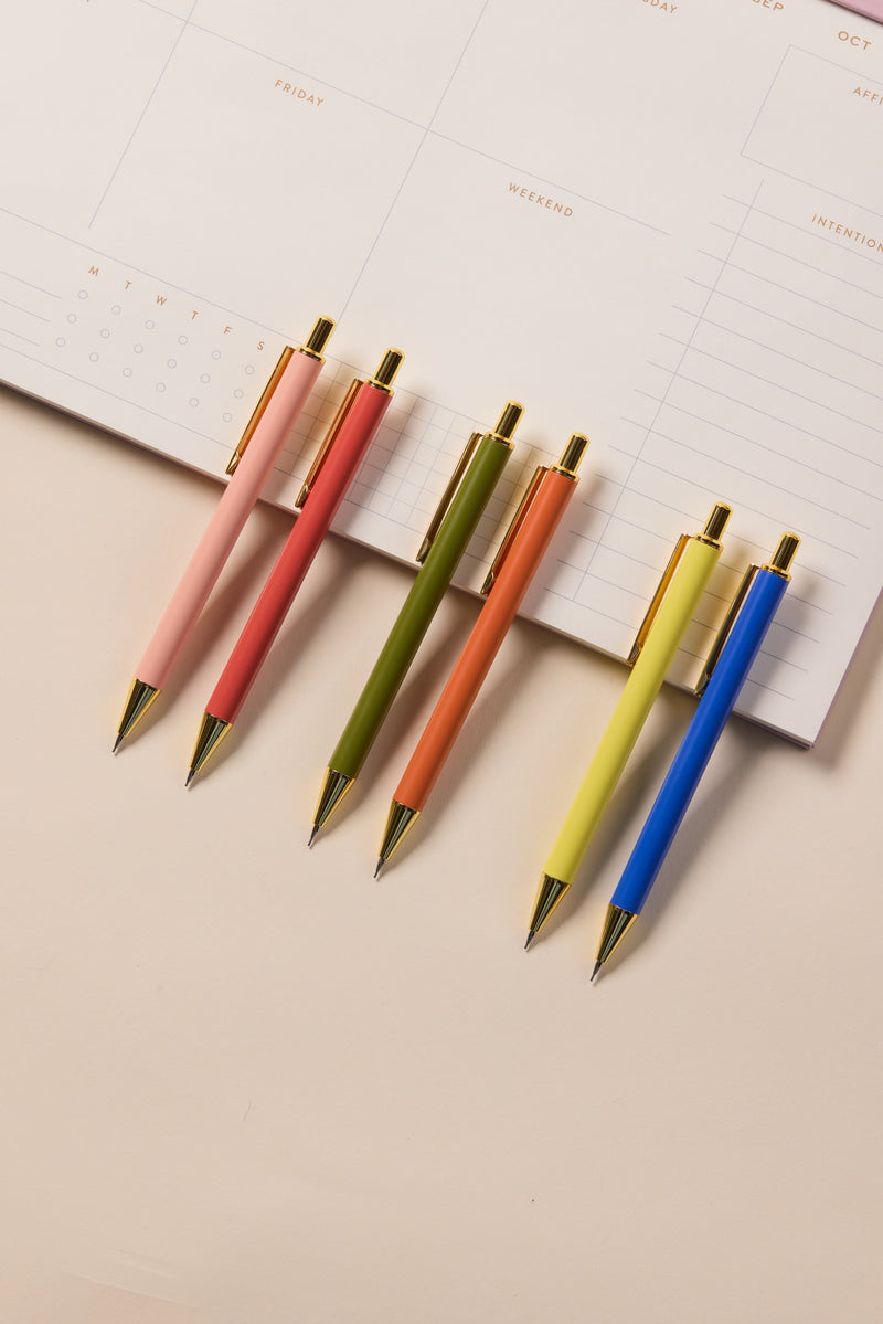 Two Mechanical Pencils (Green and Chili) are placed diagonally below an open planner with days and notes sections, providing a smooth writing experience on a light, neutral background.