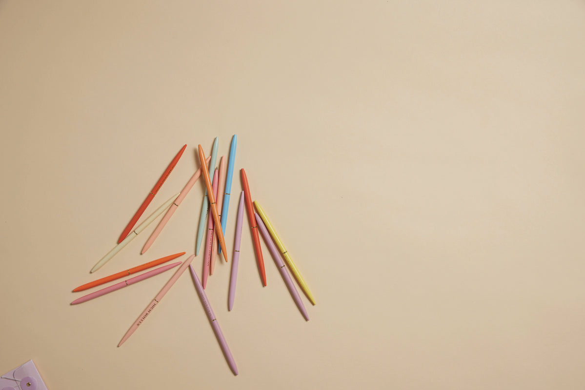 A scattered group of Slim Metal Twisty Pens - Sass Set lies on a light beige surface, with space on the right side and a small corner of paper visible in the bottom left.