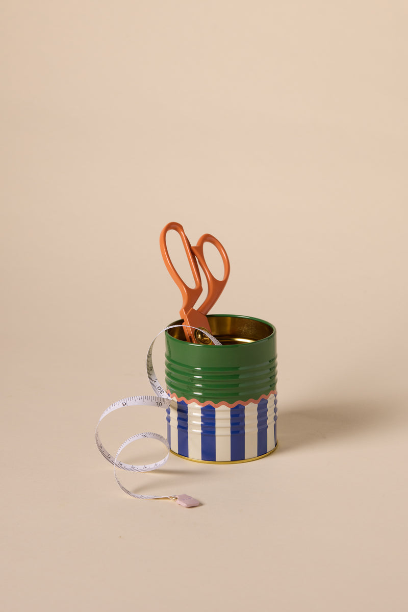 A pair of orange scissors stands upright in the A Dopo Solid & Striped Storage Tin, with a white measuring tape featuring a lavender end draped over the front, all set against a plain beige background.