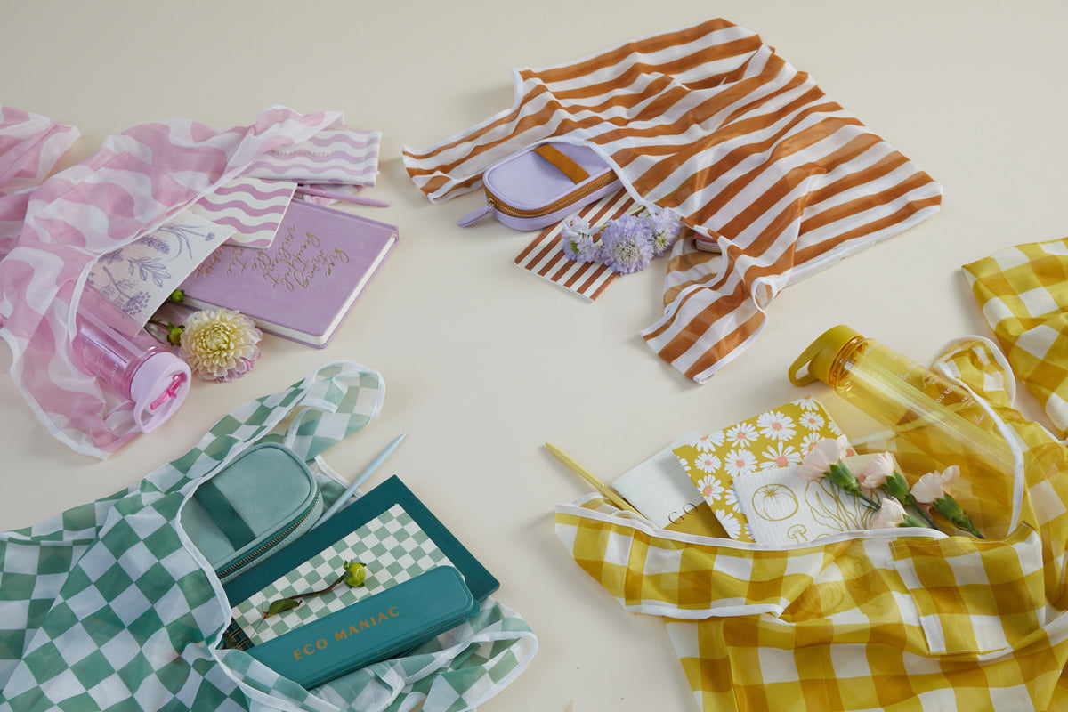 Four Reusable Market Bag - Checkered bags in pink, orange, green, and yellow hold notebooks, pens, water bottles, and flowers, all neatly arranged on a light surface.