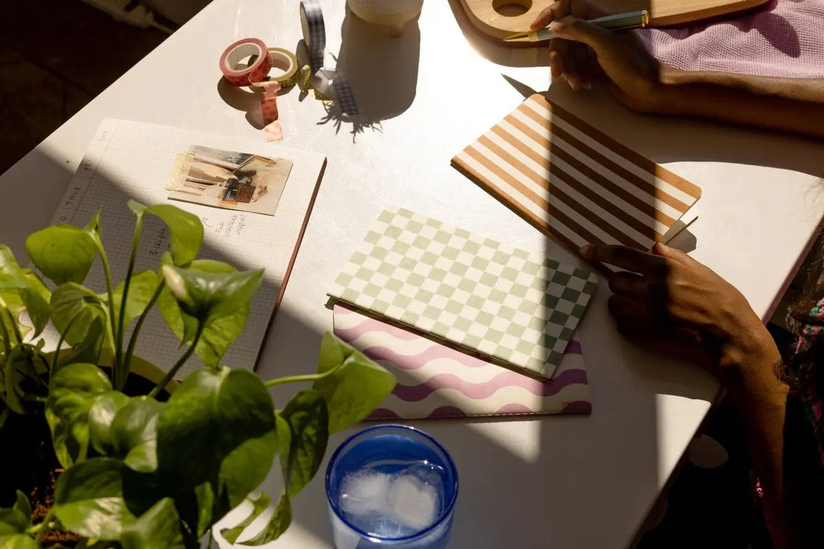 Someone writes in a Set of 3 Single Flex Undated Planners (dot, grid, sketch) on a sunlit table with patterned notebooks, washi tapes, a plant, and iced water—an ideal gift for any paper-lover.