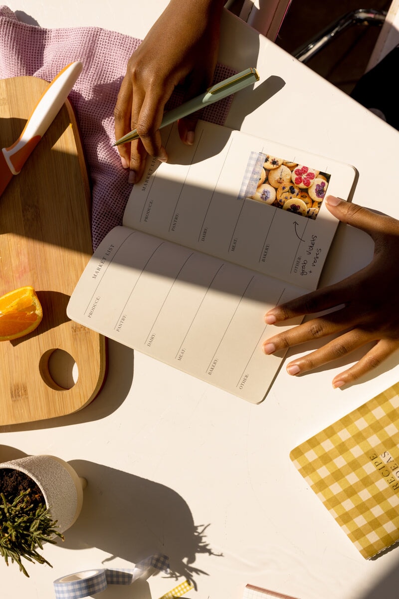 A person writes in a Set of 3 Single Flex Undated Planners - Kitchen (Meal Planner, Market List, Recipe Ideas) at a table with a checkered notebook, cutting board, orange slice, knife, and small potted plant—ideal for unique interiors.