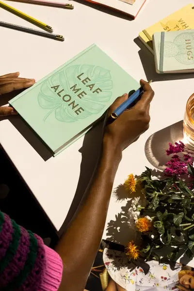 A person holds the Vintage Sass Notebook with Pen - Leaf Me Alone, featuring a mint green cover, surrounded by flowers, water, and stationery—ideal for note-taking and fans of unique design.