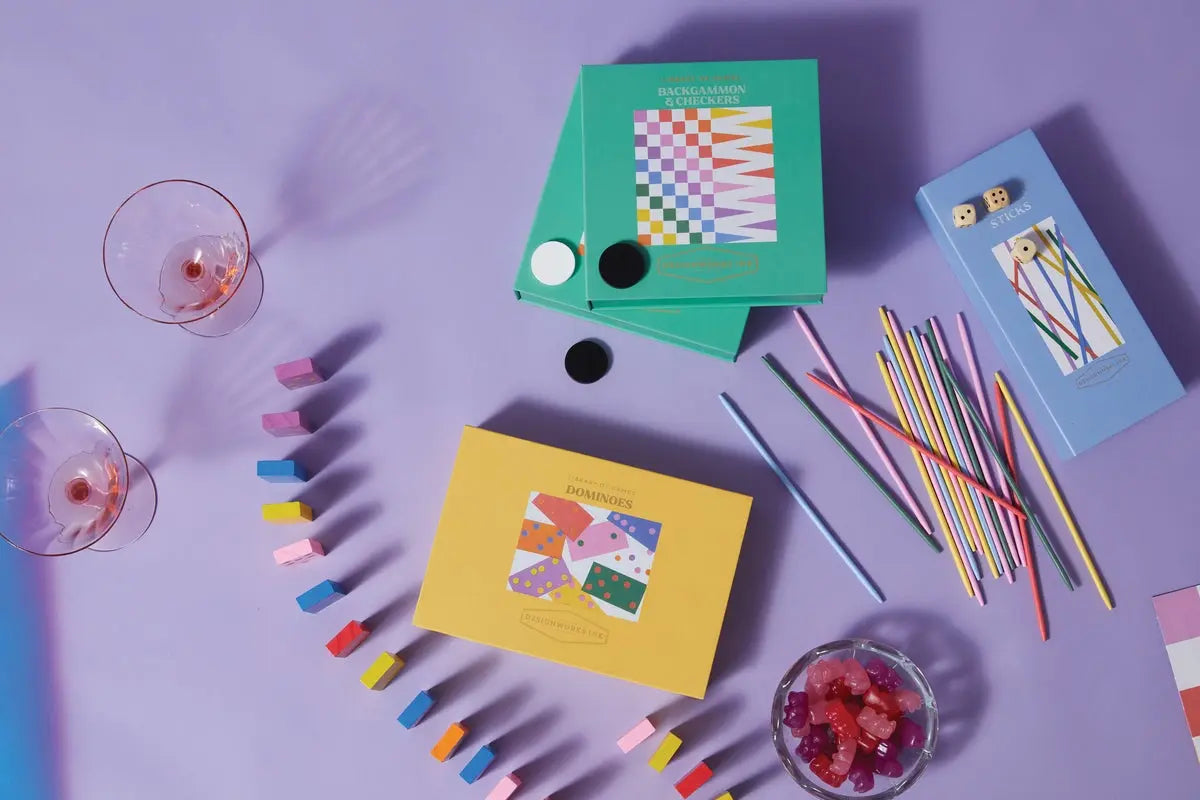 A flat lay of pastel board games, playing pieces, wine glasses with drinks, and a bowl of red gummies on a purple surface—perfect for Game Night with Timeless Classics and the Library of Games - Pick Up Sticks.