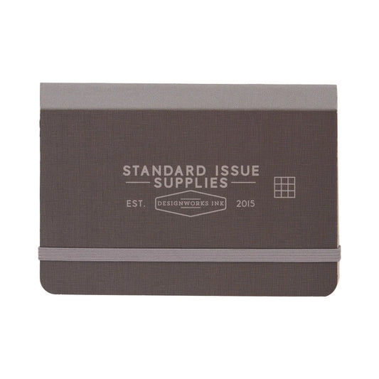 The Standard Issue Pocket Journal - Black is a compact journal with an elastic band closure, grid pages, and Standard Issue Supplies and Designworks Ink 2015 on the cover.
