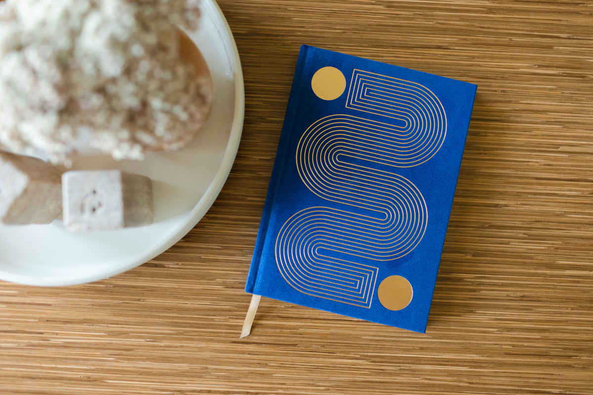The Radiant Journal - Cobalt, featuring geometric gold and white designs, rests on a wooden surface by a white tray with a potted plant and stones. This elegant lined journal is ideal for your daily notes or reflections.