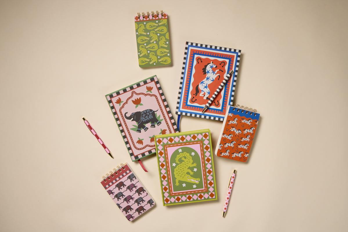 Seven vibrant notebooks with animal illustrations—including the A Dopo Elephant Twin Wire Notepad—and two patterned pens are arranged on a light beige surface. This eco-friendly stationery set features playful, bold designs perfect for creative inspiration.