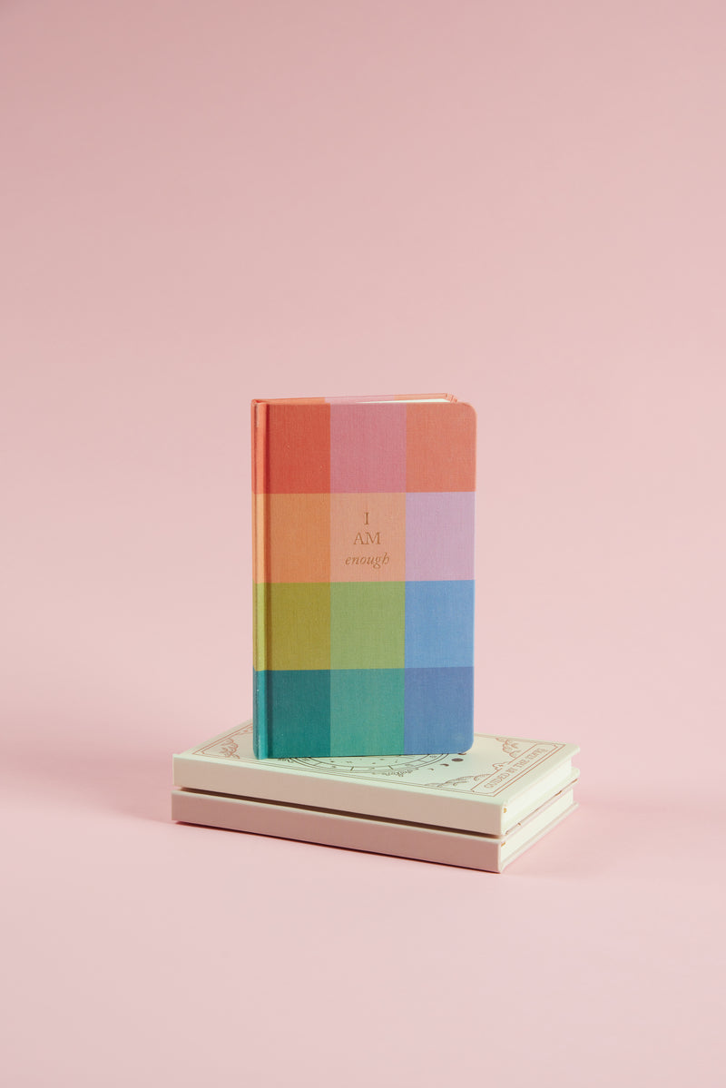 The Bookcloth Journal - Rainbow Check, featuring "I AM enough" on its cover, stands upright on two stacked white books against a pale pink background.