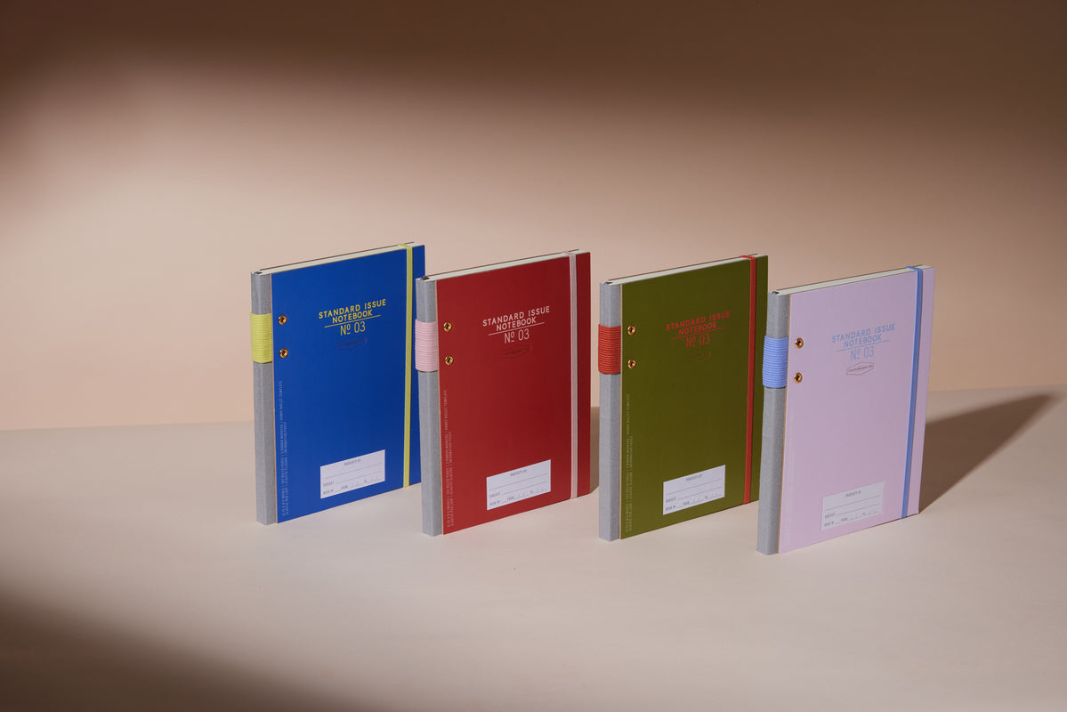Four upright Standard Issue Planner Notebook No. 03 - Rosewood + Blush notebooks stand in a row on a beige surface, each with a white spine, front label, and available in blue, red, green, or pink against a neutral background.