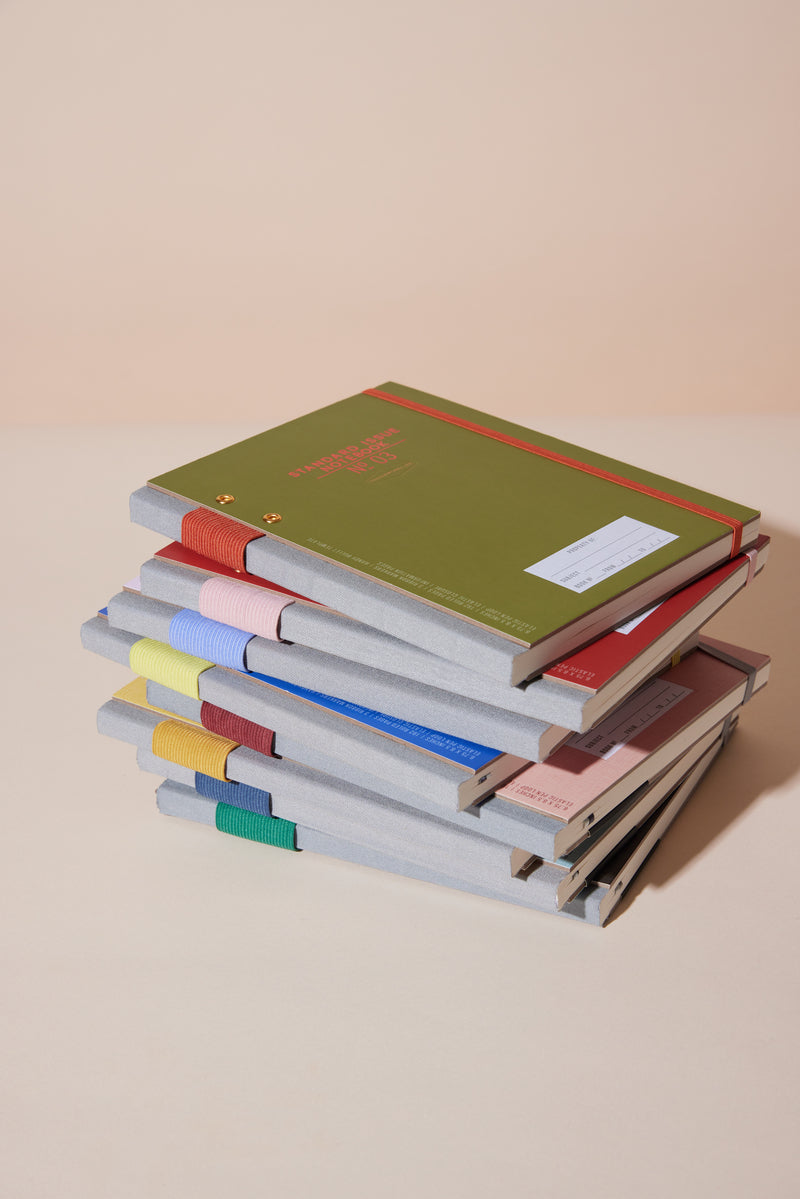 A stack of colorful hardcover notebooks with gray spines, including the Standard Issue Planner Notebook No. 03 - Green, is displayed on a beige surface. The notebooks feature varied cover colors, lined pages, and ribbon bookmarks.