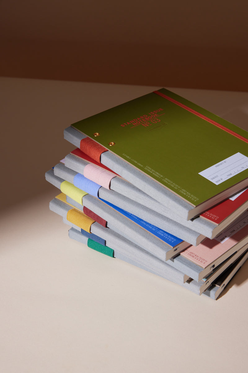 A stack of colorful notebooks, including the Standard Issue Planner Notebook No. 03 - Army Green + Chili with a textured gray spine, sits neatly arranged on a light beige surface alongside red, pink, blue, and yellow covers.