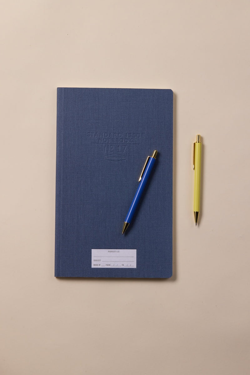 The Standard Issue Tall Notebook No.17 - Blue, known for its high-quality construction and white label, is paired with blue and yellow pens and rests on a beige surface.