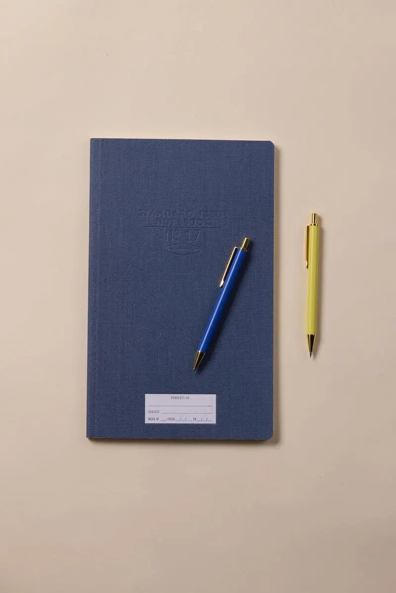 The Standard Issue Tall Notebook No.17 - Blue, known for its high-quality construction and white label, is paired with blue and yellow pens and rests on a beige surface.