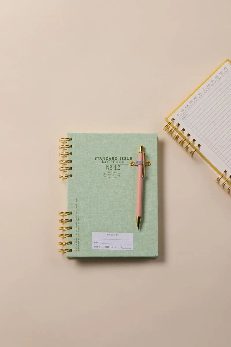The Standard Issue Notebook No.12 - Green is shown with a pink pen on a beige surface beside an open lined spiral notebook, highlighting its high-quality construction and premium writing experience.