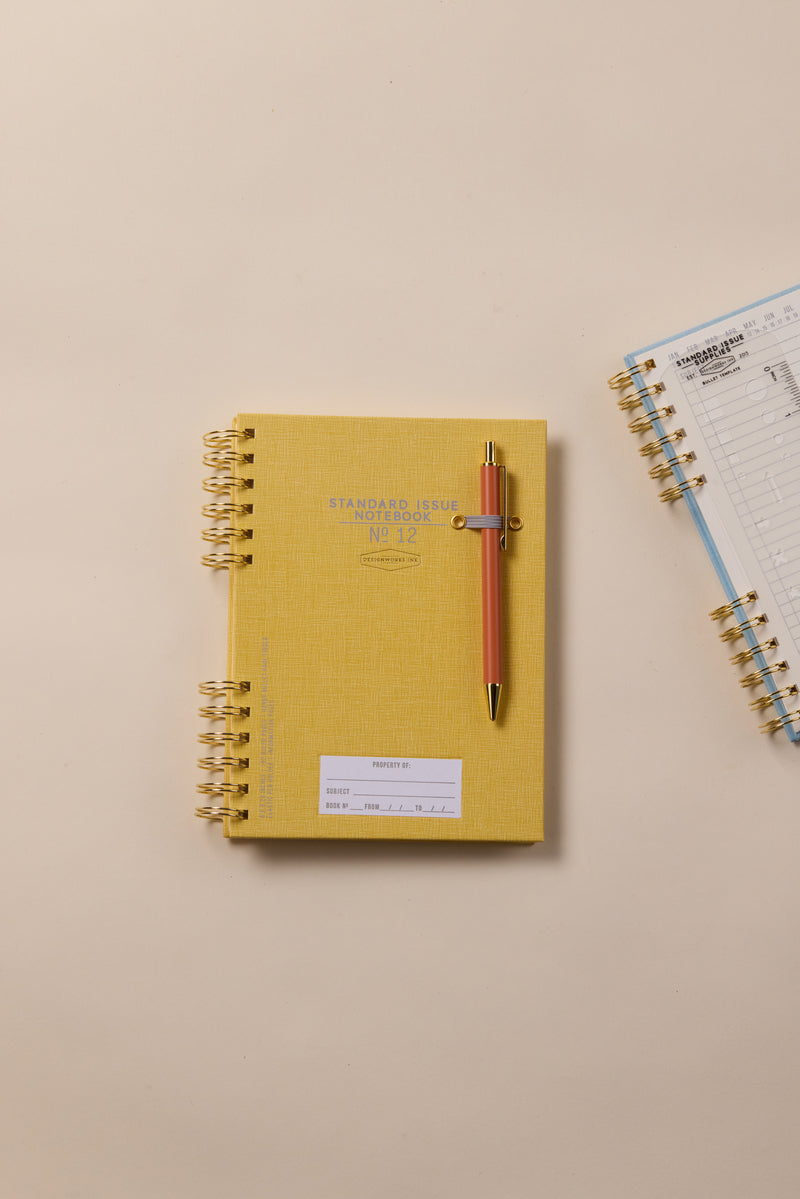 The Standard Issue Notebook No.12 - Ochre, paired with a gold and peach pen, rests on a beige surface for a premium writing experience. To its right, an open spiral-bound notebook lies nearby.