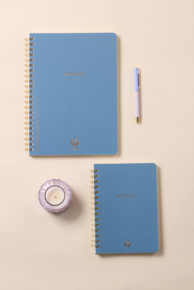 Two Textured Paper Small Twin Wire Notebooks in blue, a pastel purple and gold pen, and a lavender candle in a glass holder are neatly arranged on a beige surface.