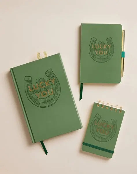 The Vintage Sass Notepad - Lucky You set includes three green notebooks with Lucky You! and a horseshoe on the cover; the largest and smallest have ribbon bookmarks, while the middle one holds a pen for retro charm.
