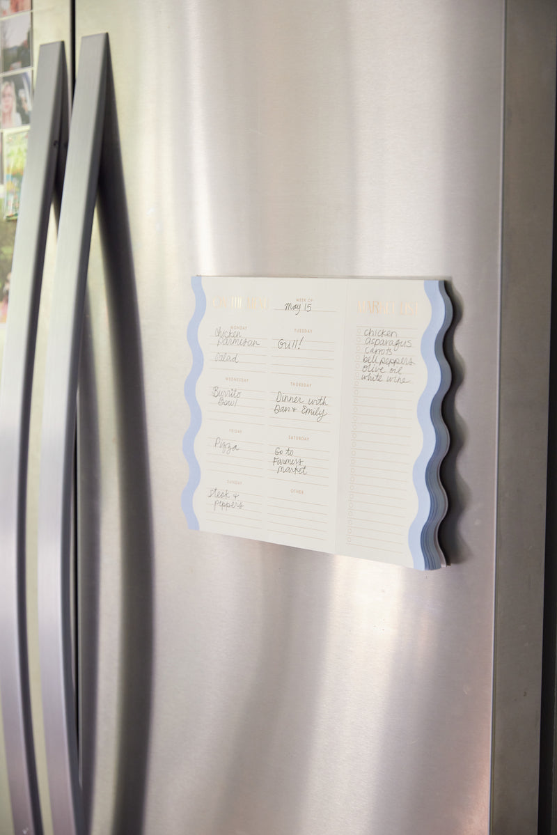 The Meal Planner Notepad with Magnets - Cloud + Cream is attached to a stainless steel fridge, open to display handwritten weekly meal and market plans beside a photo collage on the door.