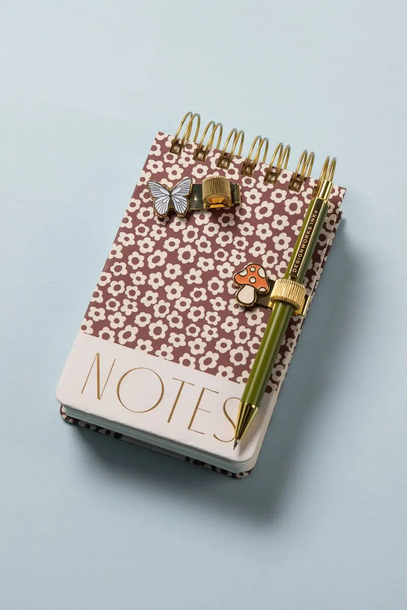 A small spiral-bound notebook with a brown floral cover, a gold pen with the Set of 2 Pen Clips - Butterfly & Mushroom, and two enamel pins—a butterfly and a mushroom—rest on a light blue surface.