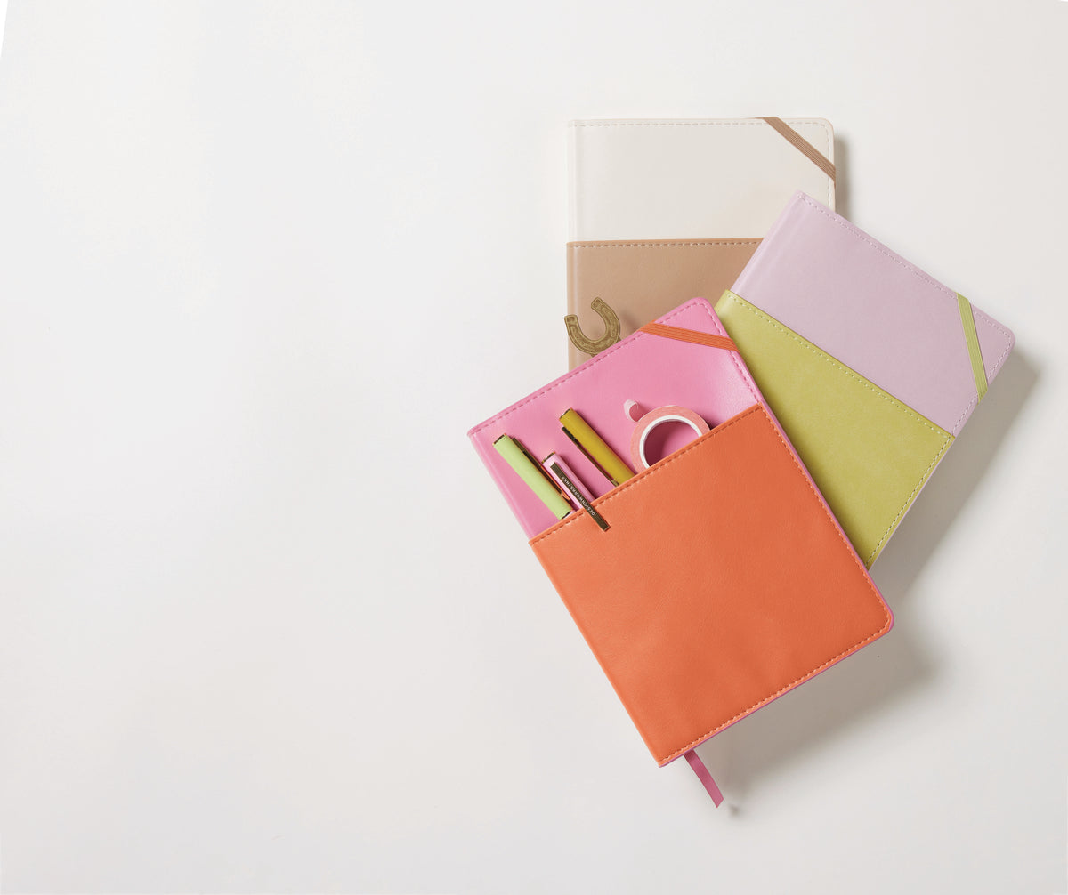 Three colorful notebooks—a Vegan Leather Pocket Journal in Pink + Chili, a white and beige notebook, and a pink journal with green accents—are arranged on a light background.