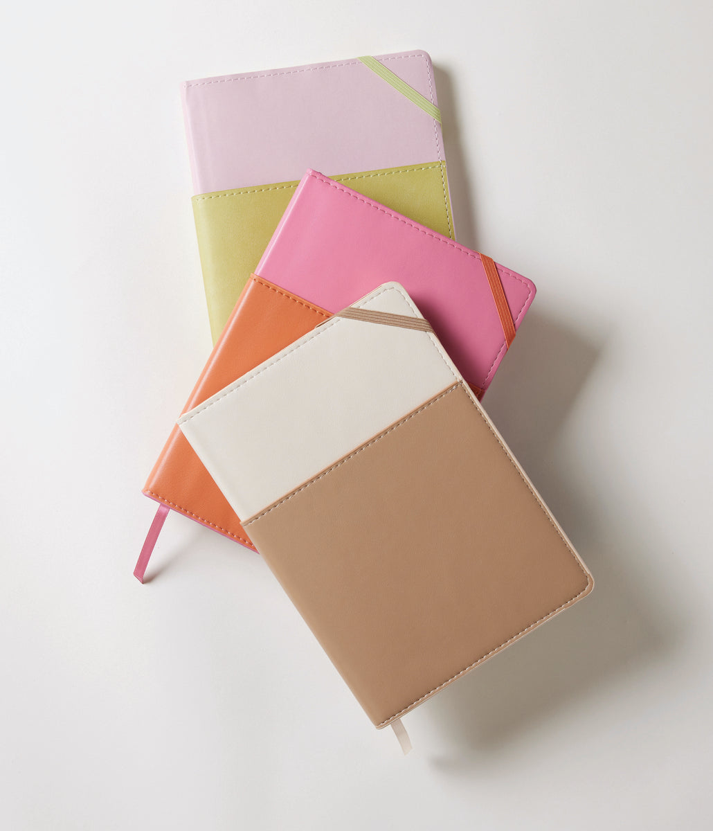 Two Vegan Leather Pocket Journals in Ivory and Oat Milk, featuring geometric designs in pastel shades and matching ribbon bookmarks, are stacked on a white surface. Each compact notebook is crafted from soft vegan leather.
