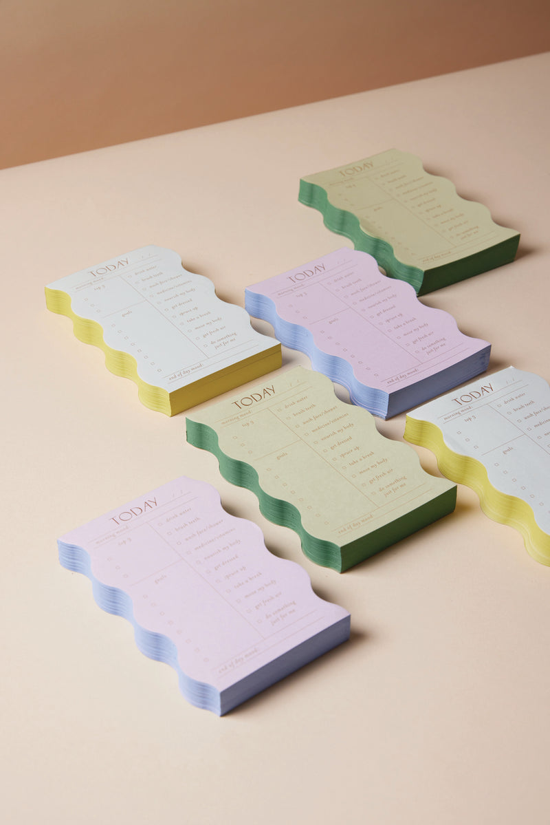 Stacks of pastel Wavy Daily Notepad - Cloud, featuring chic wavy edges and labeled TODAY, are neatly arranged on a beige surface. Each notepad includes sections for daily tasks, notes, and doubles as a self-care tracker.