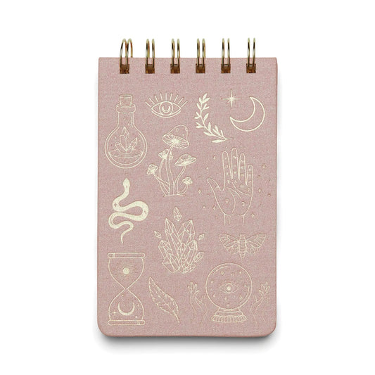 The Twin Wire Notepad - Mystic Icons is a compact, spiral-bound pink notepad featuring gold illustrations—moon, crystal ball, snake, eye, hand, plants, hourglass, and geometric shapes—on the cover. Perfect for notes on the go.