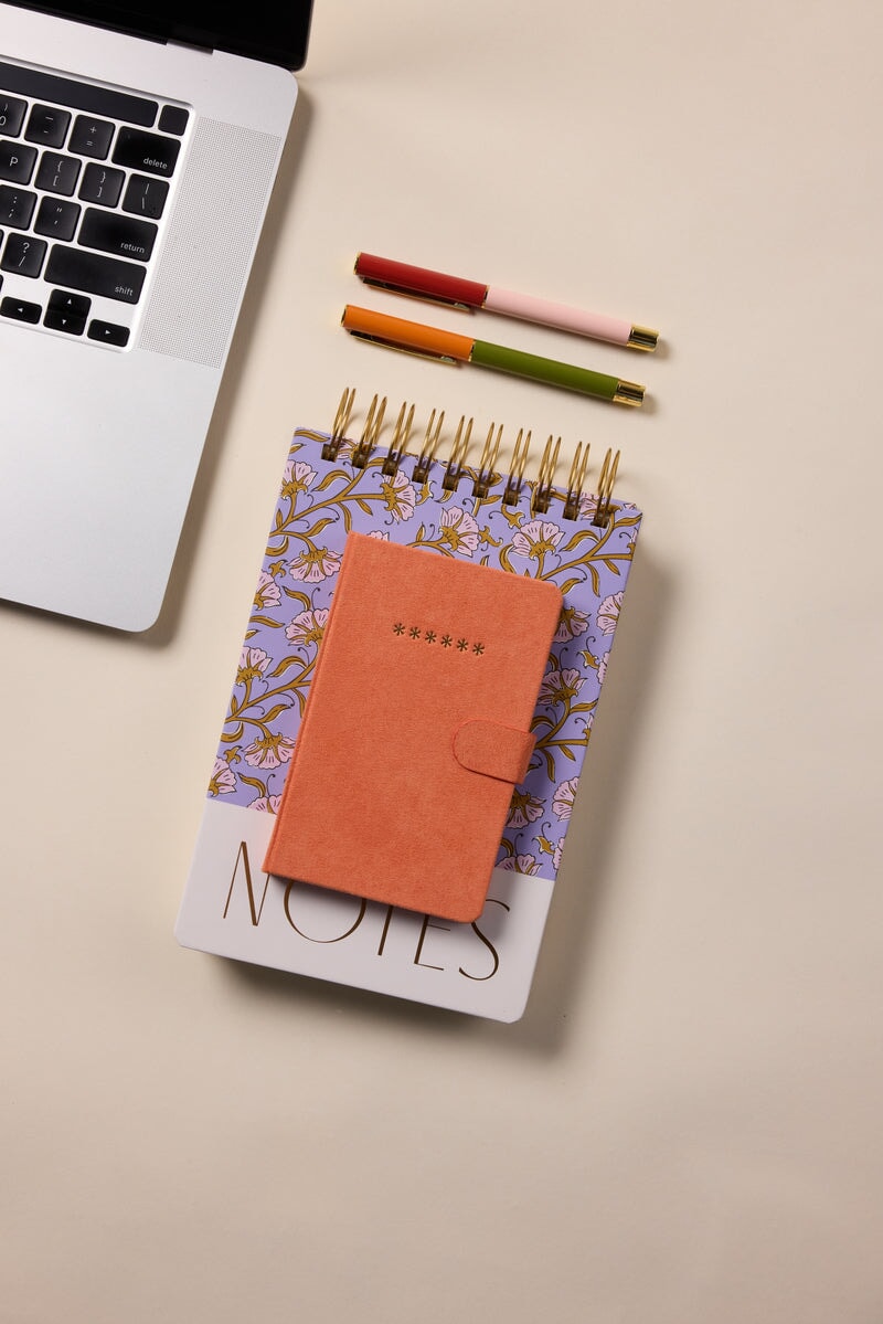A closed orange Mini Password Journal - Asterisks, a floral spiral notebook, and a NOTES notepad sit beside three pens and a laptop on a beige surface—ideal for keeping digital secrets and confidential login details safe.