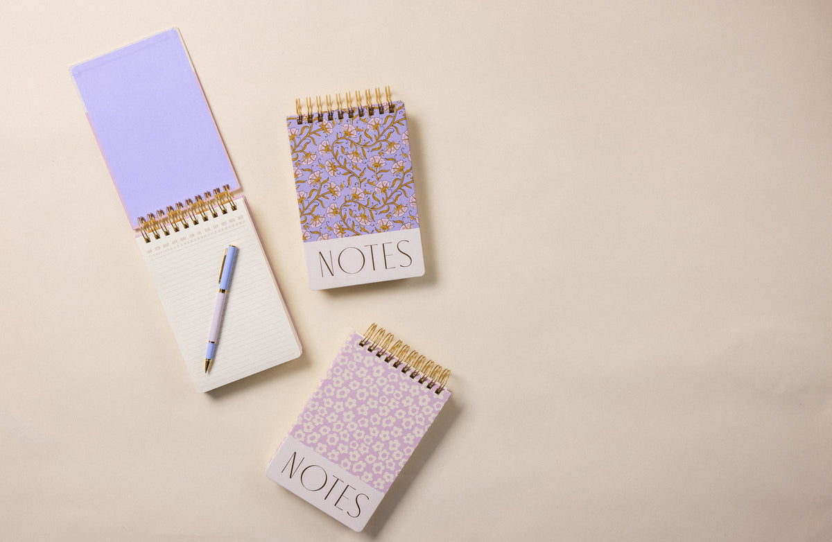 The Chunky Large Twin Wire Notepad - Lilac Groovy Floral, along with two other spiral notepads, is displayed on a beige surface; one is open with a pen resting on its pages.