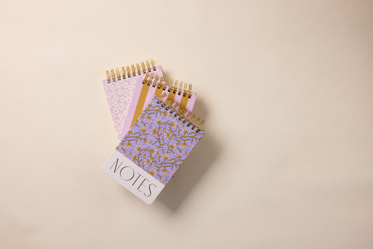 A Large Chunky Notepad - Purple Vintage Floral with a hard cover and "NOTES" written on the front rests atop two other spiral-bound, lined notepads with pastel and gold floral designs, all stacked on a beige surface.