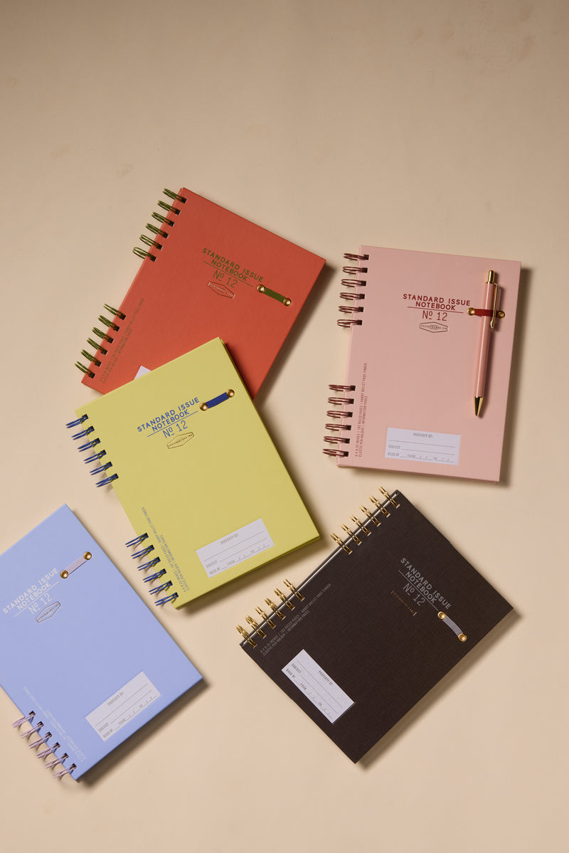 Two Standard Issue Planner Notebook No. 12 in Army Green and Chili are displayed on a beige surface, with pens clipped to their covers for a premium writing experience.