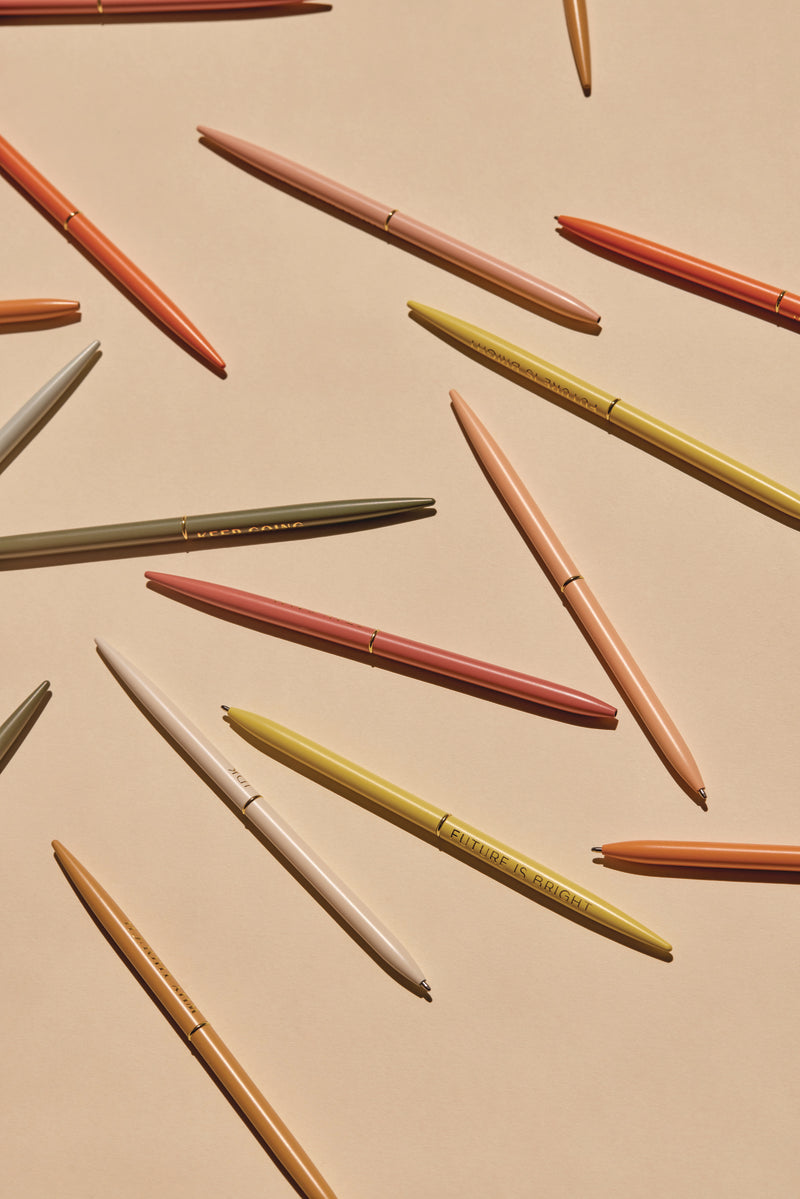 Slim Metal Twisty Pens - Positivity Set in muted shades of yellow, pink, olive, and orange are neatly arranged in a diagonal pattern across a beige background.