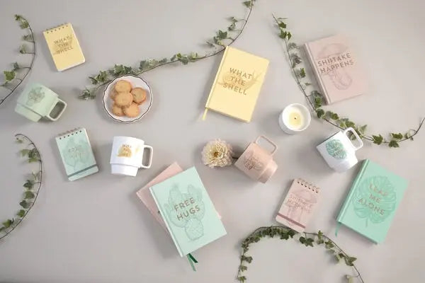 A flat lay featuring the Vintage Sass Notebook with Pen - Leaf Me Alone, mugs, spiral notepads for journaling or note-taking, a plate of cookies, a candle, and green vines arranged on a light surface.