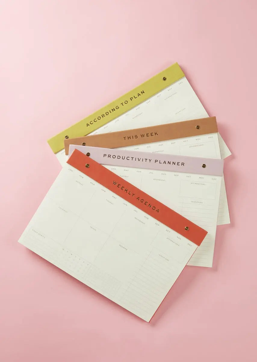 Four weekly planner pads, including the Weekly Postbound Notepad - Lilac with colored headers—According to Plan, This Week, Productivity Planner, and Weekly Agenda—are arranged on a pink background for serious organizing zen.